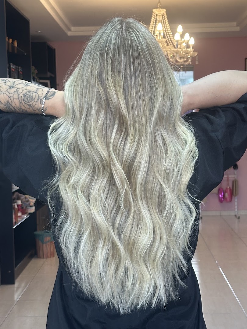 Cliente de costas com cabelo loiro em ondas longas, avental preto e tatuagem de flores no braço, lustre dourado ao fundo